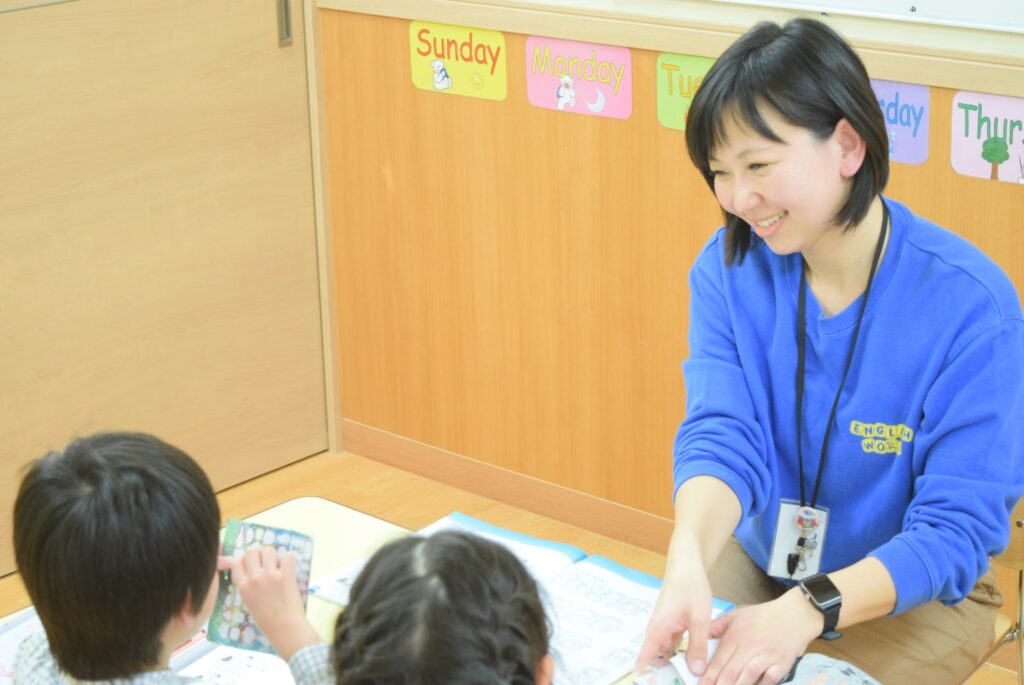 青いトレーナーを着た講師が、笑顔で子どもたちに英語のレッスンをしている様子